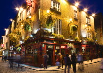 Ireland, County Dublin, Dublin City, Temple Bar Pub illuminated at night with people walking past on the cobbled streets of the area on the south bank of the Liffey River.