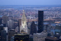 View from Empire State building over midtown skyscrapers and East River towards Queens and Long Island with Art Deco Chrysler Building and Queensboro Bridge illuminated at sunset.