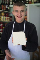 Albania, Tirane, Tirana, Young male cheese shop vendor in the Avni Rustemi Market wearing white apron and holding up block of cheese.