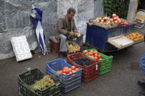 Albania, Tirane, Tirana, Fruit and vegetable street vendor seated on crate to bunch garlic behind display including grapes, oranges and onions. Part seen customer in foreground.
