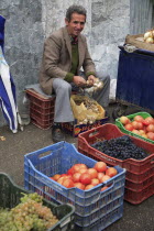 Albania, Tirane, Tirana, Fruit and vegetable street vendor in the Avni Rustemi Market seated on crate behind display including grapes and tomatoes.