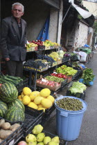 Albania, Tirane, Tirana, Grocer beside his shop front display of fruit and vegetables including melons, potatoes and grapes in the Avni Rustemi market.