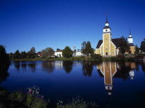 Finland, Vaasanlaani, Nykarle, Village church beside the River Lapuanjoki, Cream and white painted exterior with clock tower reflected in the water.