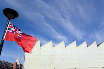 England, Hampshire, Portsmouth, Historic Naval Dockyard Red Ensign merchant marine flag flying from flagpole with Action Stations exhibition sign on building.
