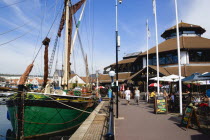 England, Hampshire, Portsmouth, Port Solent Sailing barge SB Kitty moored in the marina with people walking past a pub and restaurant on the dockside and housing beyond.