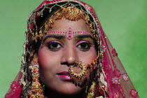 India, Uttarakhand, Hardiwar, Portrait of young woman wearing beaded pink sari and gold jewellery including nose ring, full face headshot against green background.