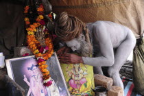 India, Uttarakhand, Hardiwar, Saddhu Baba Gajender Girri Marraj with his body covered in ash beside shrine of statue  images and offerings inside tent during Kumbh Mela festival.