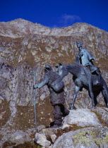Switzerland, Ticino, St Gotthard Pass, Monument to mountain guides who aided travellers across the high passes depicting figure leading horse and rider. Sheer rocky mountainside behind.
