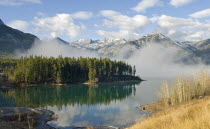 Canada, Alberta, Kananaskis, Early morning mist rising from Barrier Lake with Heart Mountain and the Rockies in the background, reflection of pine trees in lake.