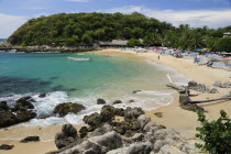 Mexico, Oaxaca, Puerto Escondido, View onto Playa Manzanillo beach with rocks and driftwood in foreground, tourist boats, people and tree covered headland beyond.