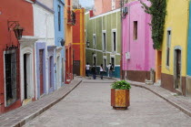 Mexico, Bajio, Guanajuato, Paved street lined by colourfully painted houses, group of four people walking at far end and container of geraniums in centre foreground.