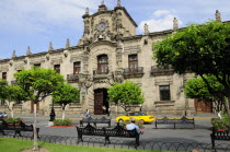 Mexico, Jalisco, Guadalajara, Palacio Gobierno, the Government Palace.  Exterior facade from Plaza de Armas with passing car, trees and man seated on bench in foreground.