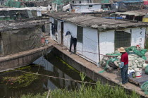 China, Jiangsu, Qidong, Man in a red shirt and straw hat fishing with a rod and line off his boat moored in a canal. Pair of black trousers hanging on a line against a white background; Many poor land...