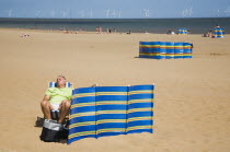 England, Lincolnshire, Skegness, Man sleeps in sunshine behind striped windbreak with Lincs Wind Farm offshore behind.