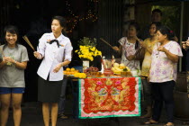 Thailand, Bangkok, Woman in military uniform holding joss sticks beside offerings on decorated table celebrating local temple parade.