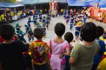 Thailand, Bangkok, Thai children watch dance troupe at local temple from elevated stage.