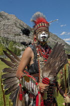 Canada, Alberta, Waterton Lakes National Park, Blackfoot head dancer Aryson Black Plume in full regalia and face paint at the Blackfoot Arts & Heritage Festival Pow Wow organized by Parks Canada and the Blackfoot Canadian Cultural Society, Headdress with arrow and black plume, Feather fan and bustle, Rocky Mountains blue sky with white clouds, red; blue; green; dominant red; UNESCO World Heritage Site.