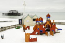 England, East Sussex, Brighton, West Pier during winter with snow on the beach and childrens play area in the foreground.