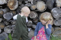 Kids, Outdoor, 5 year old Eva and Oscar aged two, playing beside a log pile with Eva using bark as pretend mobile phone.