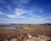 France, Brittany, Cotes d'Amor, Cote de Granit Rose, rocky shoreline with view towards ile grand on the skyline. cirrus clouds in blue sky above.