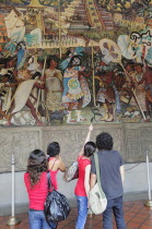 Mexico, Federal District, Mexico City, Group of visitors looking at mural by Diego Rivera depicting life before the Conquest in the Palacio Nacional.