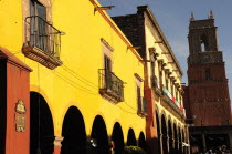 Mexico, Bajio, San Miguel de Allende, El Jardin, Yellow facade of colonial mansion and arcades with clock tower beyond.