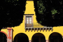 Mexico, Bajio, San Miguel de Allende, El Jardin, Detail of yellow painted facade of colonial mansion with French window and balcony, part framed by trees in foreground.