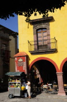 Mexico, Bajio, San Miguel de Allende, El Jardin, Part view of yellow painted facade of colonial mansion with French window and balcony, with fruit juice vendor in street below.