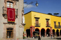 Mexico, Bajio, San Miguel de Allende, El Jardin, Part view of Museo Casa de Allende and yellow painted arcades, people and fruit juice vendor.