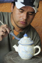 Mexico, Puebla, Talavera ceramic artist at Armando Gallery applying blue detail to raised design on teapot.