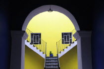 Mexico, Puebla, Looking into inner courtyard through archway in shadow towards yellow painted wall with blue and white tiled steps, plant in blue pot and pair of square, recessed windows.