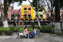 Mexico, Puebla, Two young women chatting on stone bench in the Zocalo with colourful building facades hung with Mexican flags and line of sun umbrellas and outside cafe tables beyond area of grass beh...