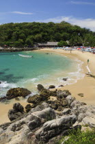 Mexico, Oaxaca, Puerto Escondido, View onto Playa Manzanillo beach with rocks in foreground and tourist boats, people and tree covered headland beyond.