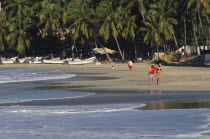 Mexico, Oaxaca, Puerto Escondido, Playa Marinera with couple walking along shore, line of boats pulled up onto sand and overhanging palms beyond.