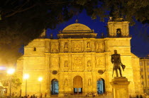 Mexico, Oaxaca, Baroque exterior facade of cathedral at night part framed by tree branches with people and statue in foreground.