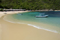 Mexico, Oaxaca, Huatulco, Playa La Entrega, Stretch of sandy beach lined with thatched open fronted restaurants, with tour boat in shallow water in foreground.