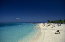 West Indies, Jamaica, Montego Bay, sandy beach beside aquamarine water with people sunbathing and in the sea  with low flying aeroplane over the water.