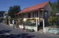West Indies, Jamaica, Port Antonio, roadside housing with veranda and walled garden with boy and young girl cycling past.