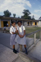 West Indies, Jamaica, Two schoolgirls in checked dresses in playground in front of school buildings.