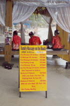 Thailand, Koh Sumui, Chaweng beach, beach massage with three women wearing red sat on benches giving massage with a sign displaying prices for various treatments in the foreground.