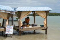 Thailand, Koh Sumui, Chaweng beach, beach massage on a four poster bench over the sea with a woman giving a man a massage with a sign displying prices for various treatments.
