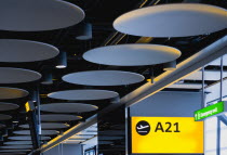 England, London, Heathrow Airport Terminal 5 disc ceiling in departures zone with passengers waiting in seating area at gate.