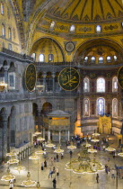 Turkey, Istanbul, Sultanahmet, Haghia Sophia Sighseeing tourists beneath the dome with murals and chandeliers in the Nave of the Cathedral with calligraphic roundels of Arabic Koran texts abobe the Lo...