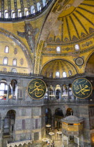 Turkey, Istanbul, Sultanahmet, Haghia Sophia Sighseeing tourists beneath the dome with murals and chandeliers in the Nave of the Cathedral with calligraphic roundels of Arabic Koran texts abobe the Lo...