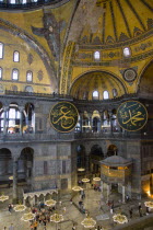 Turkey, Istanbul, Sultanahmet, Haghia Sophia Sighseeing tourists beneath the dome with murals and chandeliers in the Nave of the Cathedral with calligraphic roundels of Arabic Koran texts abobe the Lo...