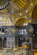 Turkey, Istanbul, Sultanahmet, Haghia Sophia Sighseeing tourists beneath the dome with murals and chandeliers in the Nave of the Cathedral with calligraphic roundels of Arabic Koran texts abobe the Lo...