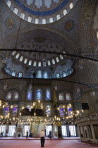 Turkey, Istanbul, Sultanahmet Camii, The Blue Mosque interior with people at prayer beneath chandeliers and decorated domes above and stained glass windows beyond.