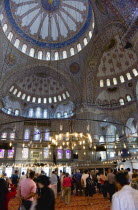 Turkey, Istanbul, Sultanahmet Camii, The Blue Mosque interior with sightseeing tourists by a chandelier below the decorated domes with stained glass windows beyond.