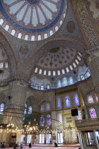 Turkey, Istanbul, Sultanahmet Camii, The Blue Mosque interior with people at prayer beneath chandeliers and decorated domes above and stained glass windows beyond.
