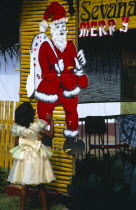 Sri Lanka, Christianity, little girl in yellow party dress looking up at large cut-out of father christmas pinned to yellow painted frame.